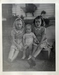 Two Little Girls Sitting On a Sidewalk on Either Side of a Baby