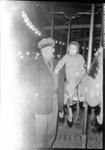 Man Standing Next to Child on Carousel by Fred A. Blocker